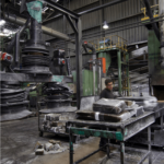 A worker operates amidst machinery in an industrial factory setting, with various materials and packaged goods on a conveyor belt and surrounding workspaces.